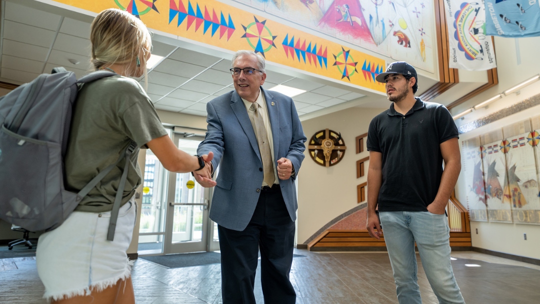 A man in a gray suit jacket and yellow tie shakes hand with a blonde student.