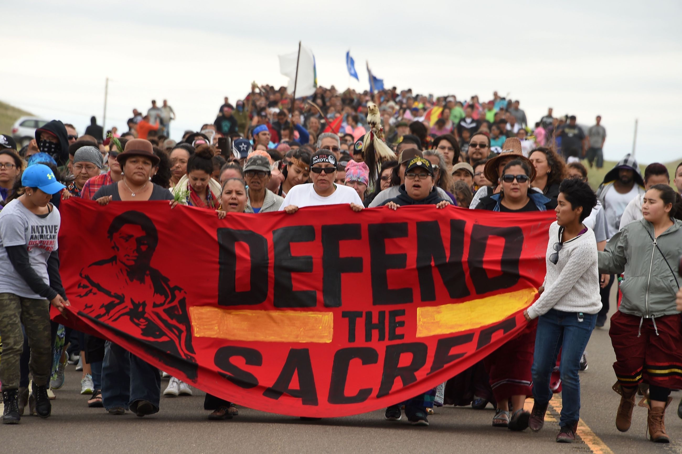 A large group of people march and carry a sign that says 'defend the sacred'
