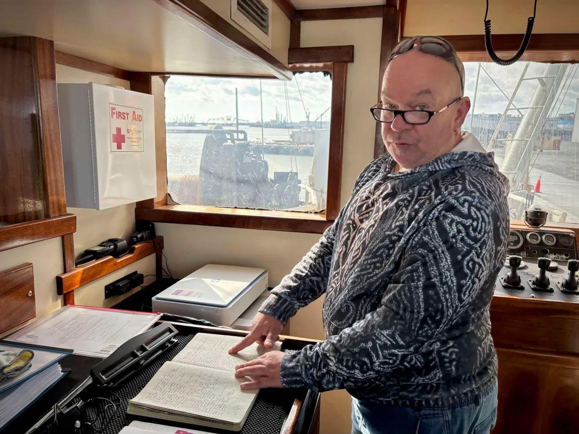 A bald man in glasses and a gray sweatshirt stands in the cabin of a boat