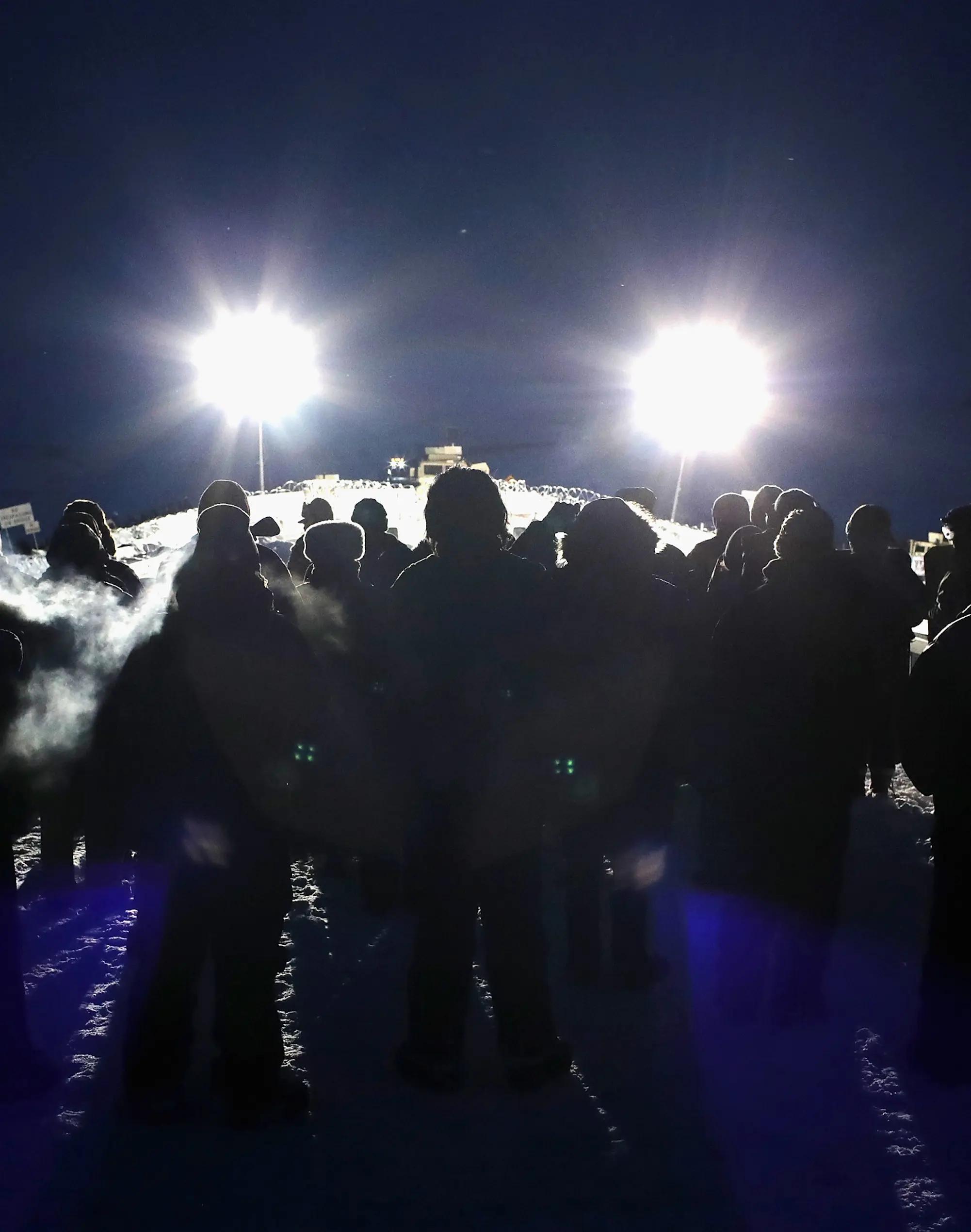 A group of people silhouetted against bright lights face off with police or security forces at night.
